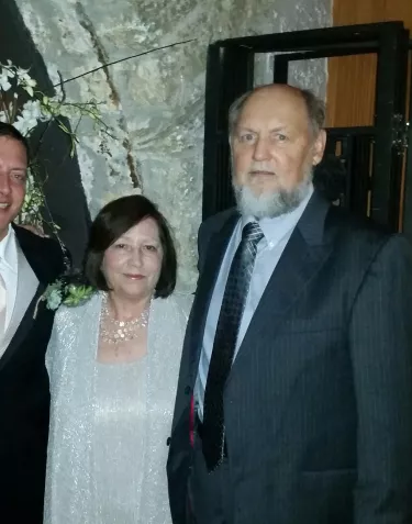 Four people dressed formally pose at what appears to be a special event or celebration. The setting includes decorative branches with white flowers. The woman on the left wears a sparkly navy dress, the man next to her wears a black suit with white tie, and the couple on the right wear silver/gray formal attire.