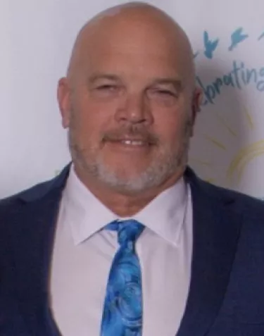 A professional headshot of a person wearing a navy suit jacket, light blue dress shirt, and blue patterned tie. They have a bald head and well-groomed gray beard, and are smiling warmly at the camera against a light background with some decorative elements.