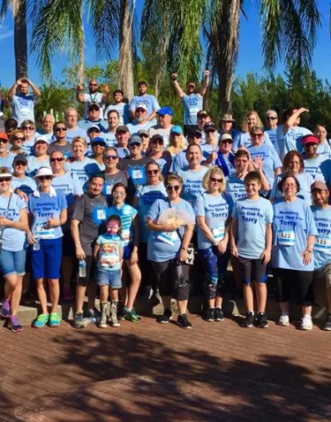 A large group of people wearing light blue t-shirts with "Supporting Terry" text gather outdoors for a group photo. The backdrop includes palm trees and a brick pathway on a sunny day. The group appears to be participating in a prostate cancer awareness or fundraising event.