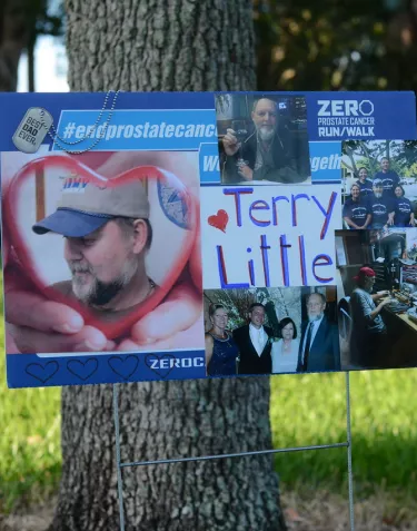 A yard sign for ZERO Prostate Cancer Run/Walk features a collage of photos of Terry Little, including personal photos and event pictures. The sign includes the ZERO organization logo and is displayed on a lawn with a tree trunk visible in the background.