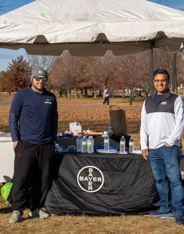 Two men standing under a white tent in a park setting. One is wearing a navy blue shirt and a beanie, and the other is wearing a white and black shirt. A table with a "Bayer" logo is in front of them, displaying water bottles and snacks.