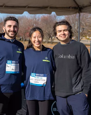 Three people standing together under a tent in a park. Two are wearing navy blue shirts with event badges, and one is wearing a black Columbia sweatshirt. They are smiling and appear to be participating in a prostate cancer awareness event.