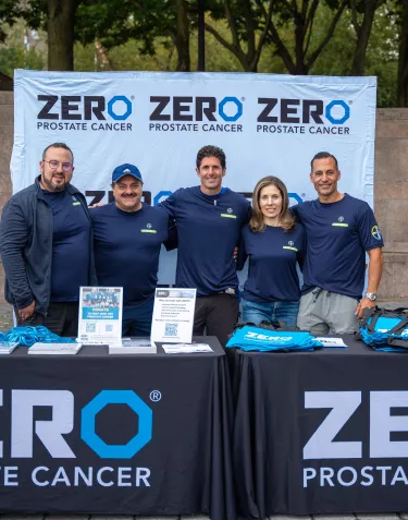 A group of five people standing behind a table with "ZERO Prostate Cancer" banners. They are smiling and wearing matching navy blue shirts. The table is covered with promotional materials and items related to the cause.