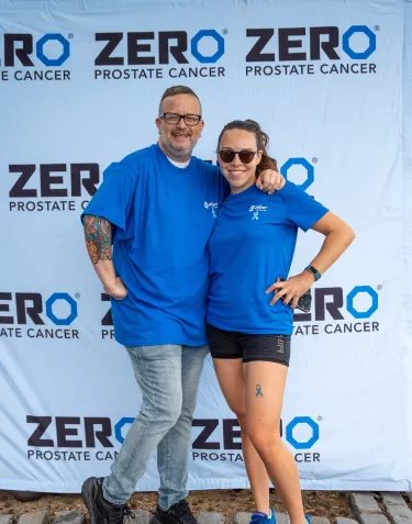 Two people smiling and posing in front of a "ZERO Prostate Cancer" banner. They are wearing blue Pfizer shirts, with one person having a tattoo on their leg.