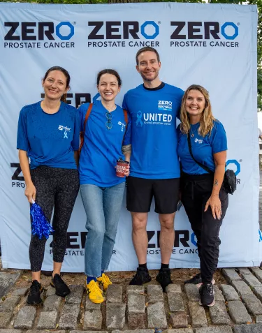 Four people standing together, smiling in front of a "ZERO Prostate Cancer" banner. They are wearing blue shirts, and the setting appears to be a park.