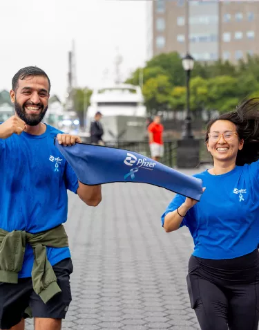 A man and a woman running joyfully, holding a blue Pfizer banner. They are wearing blue shirts, and the background shows a waterfront area with buildings and trees.