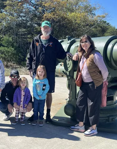 A group of people posing together next to a large green military cannon or artillery piece displayed outdoors. The group includes adults and children dressed casually, with trees and natural scenery visible in the background on a sunny day.