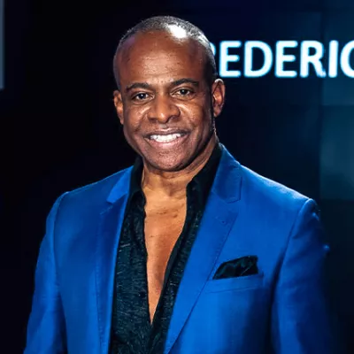 A middle aged Black man wearing a dark blue shirt with open buttons to his mid-chest and a royal blue jacket. 