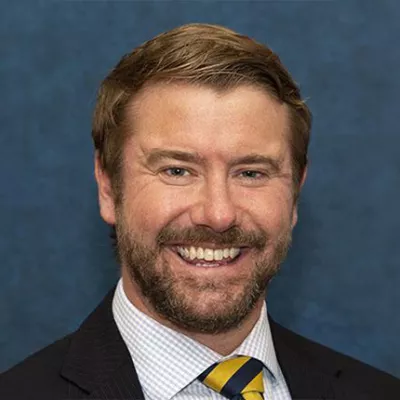 A middle aged white man with light brown hair and a beard smiling against a blue background