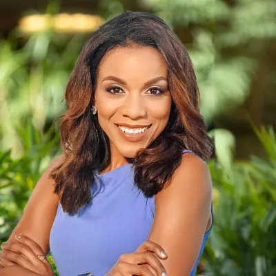 A Black woman in a purple top standing with her arms crossed and smiling in front of green foliage