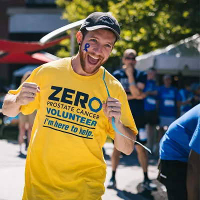 An energetic volunteer wearing a yellow ZERO Prostate Cancer Volunteer t-shirt with "I'm here to help" text smiles broadly at the camera on a sunny day. He has a blue ribbon painted on his face, with other volunteers visible in the background.