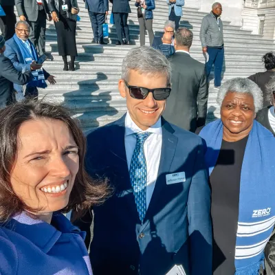 A group of people smiling on the steps of a large building, with some wearing blue scarves that say "ZERO Prostate Cancer." The setting appears to be a formal gathering or event.