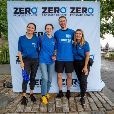 Four people standing together, smiling in front of a "ZERO Prostate Cancer" banner. They are wearing blue shirts, and the setting appears to be a park.