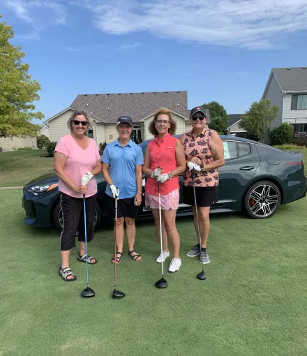 Four women in golf attire standing with drivers in front of a luxury gray vehicle on a golf course