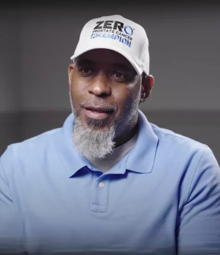 A black man with a gray beard against a gray background, wearing a light blue polo and a white hat that says ZERO Champions