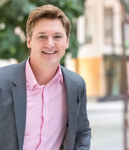 A blonde man wearing a pink shirt and gray jacket