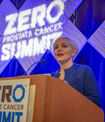 Blonde woman with very short hair standing behind a podium that has a sign saying "ZERO Prostate Cancer Summit"