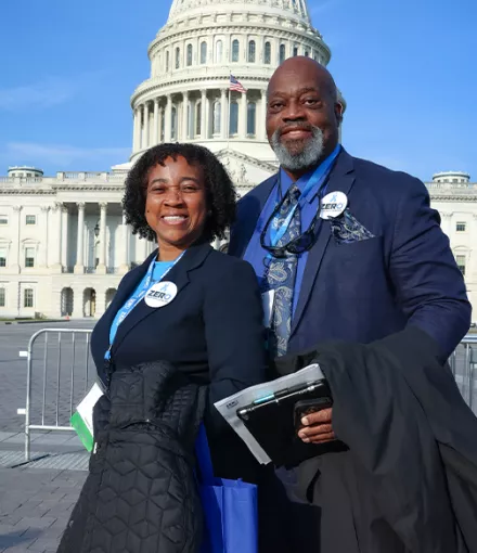 Two advocates wearing ZERO badges stand smiling in front of the U.S. Capitol building in Washington, D.C. on a sunny day. They are dressed professionally, with one person holding what appears to be documents or materials.