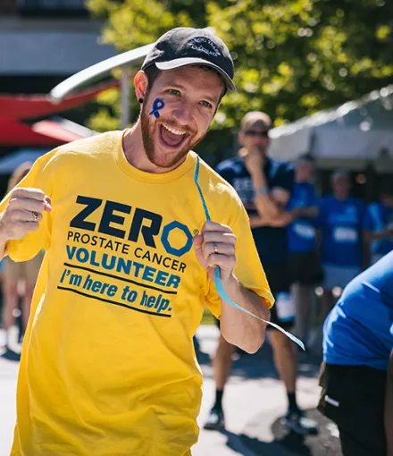 An energetic volunteer wearing a yellow ZERO Prostate Cancer Volunteer t-shirt with "I'm here to help" text smiles broadly at the camera on a sunny day. He has a blue ribbon painted on his face, with other volunteers visible in the background.