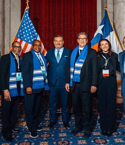 A group of seven people stands together indoors, dressed in formal attire. Some are wearing blue scarves with "ZERO" logos. They are positioned in front of American and Texas flags, with a red curtain in the background.