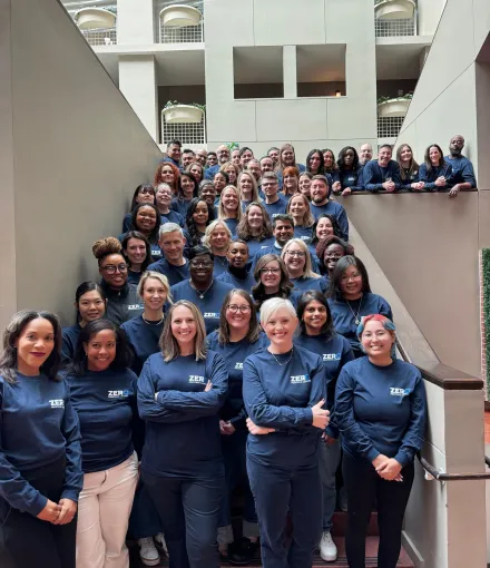 A large group of people standing on a staircase, all wearing matching navy blue shirts with the text "Bigger, Bolder, Zero" printed on them. They are smiling and posing for the photo in a well-lit indoor setting.