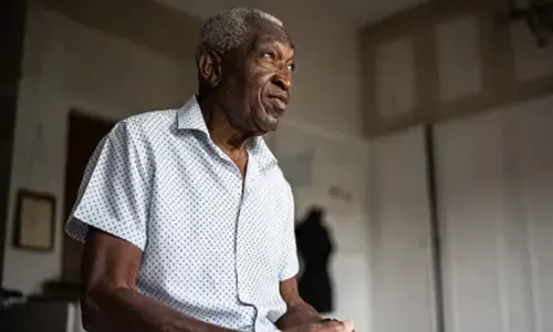 A Black man sitting on the edge of his bed with his hands together