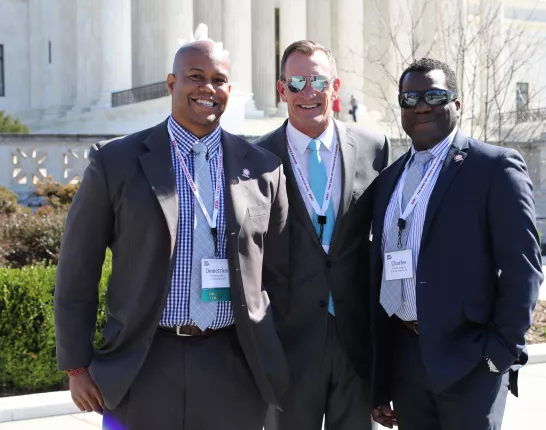 Three sharply dressed men in suits wearing lanyards and stand in front of a formal marble building