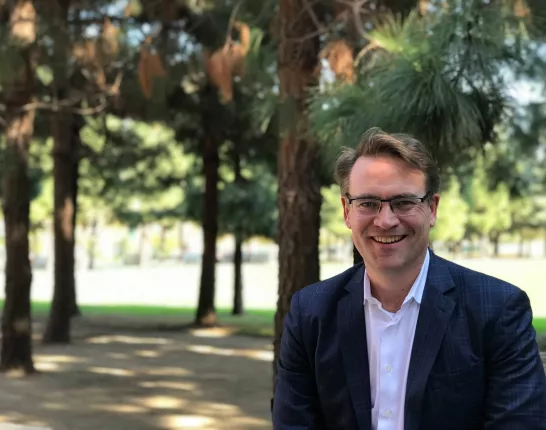 A man wearing smart looking glasses in a suit without a tie sits in shady trees out in the sun