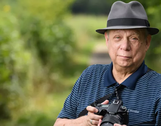 Older man wearing gray fedora holding a dslr camera on a forest path