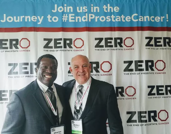 Mike Crosby and Chas Rodgers stand in front of a journey to end prostate cancer step and repeat