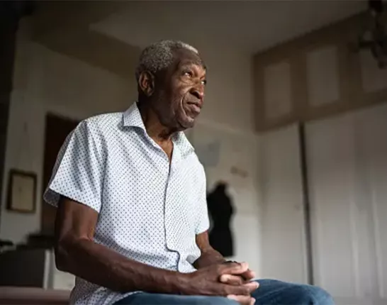 A Black man sitting on the edge of his bed with his hands together
