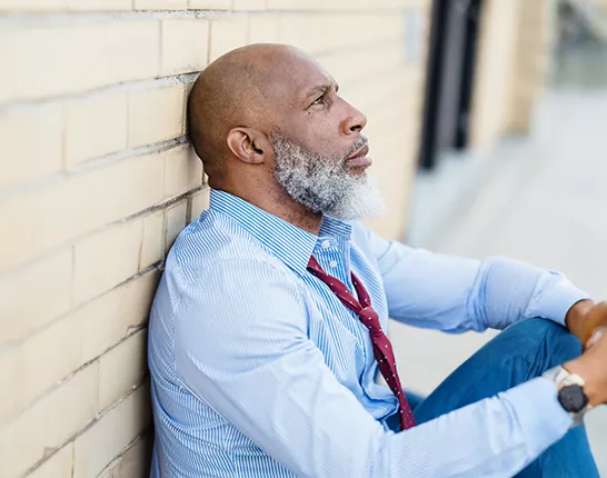 Black man sitting on the ground against a brick wall looking distressed
