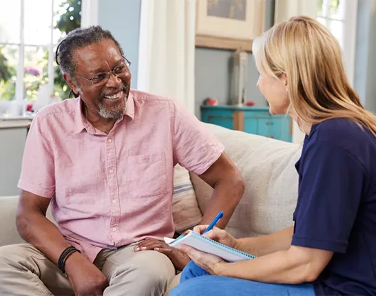 An older Black man sitting on a couch with a woman who is holding a clipboard