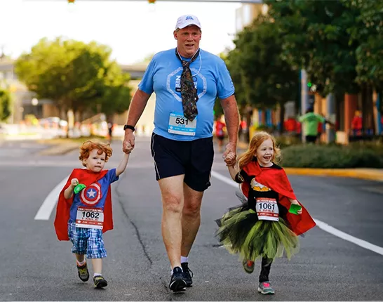 Man holding hands with his two children who are wearing superhero costumes at a Run/Walk