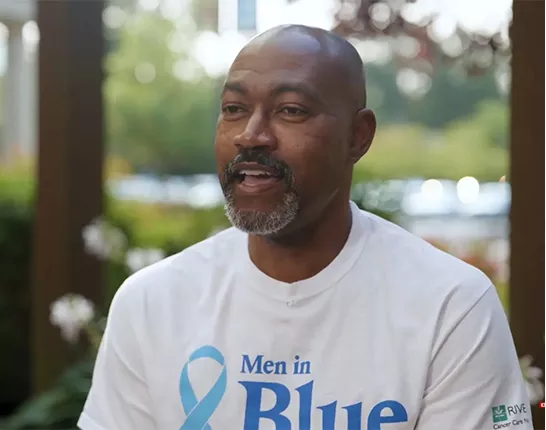 Terrance Gist, a prostate cancer patient in Williamsburg, VA, sitting down wearing a white shirt that has a blue prostate cancer ribbon that says "Men in Blue"