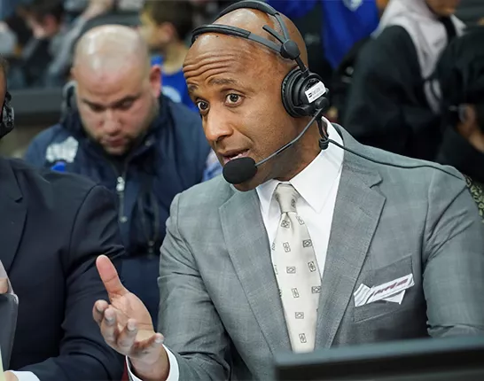 A Black man (Brian Custer) wearing a suite and a headset as an announcer at a sporting event