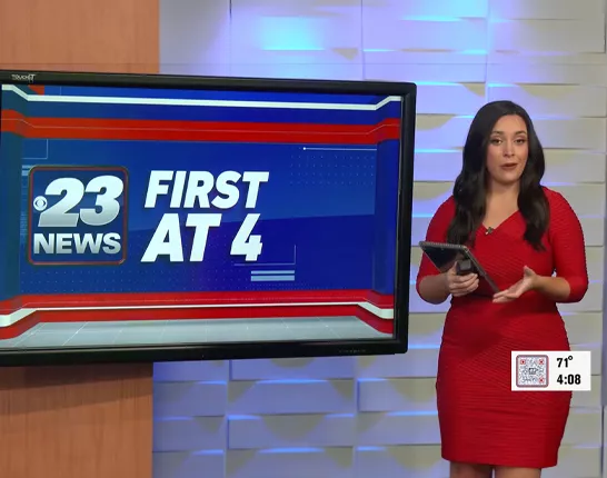 A news anchor wearing a red dress standing next to a television screen that says "23 News First at 4"