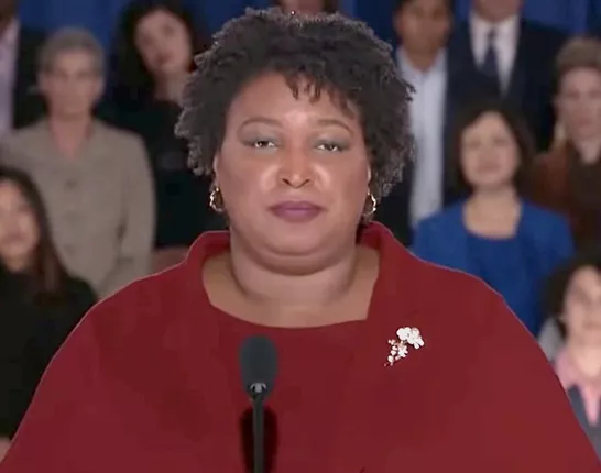 Stacy Abrams speaking at a microphone wearing a classic red sweater