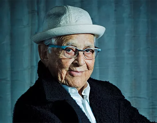 TV writer and personality, Norman Lear, wearing a white hat and dark jacket while sitting in front of a gloomy blue background