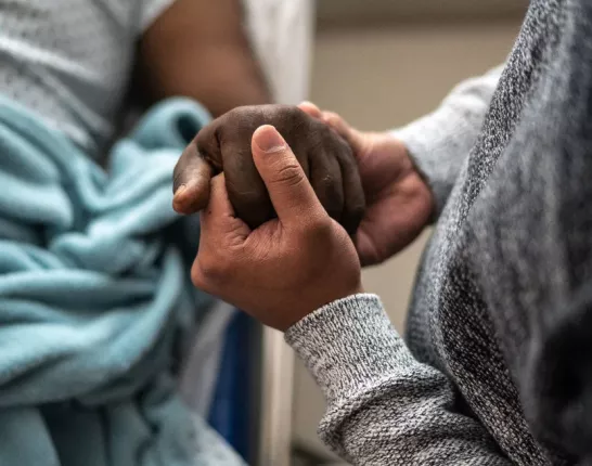 Two black people holding hands; one of them is in a hospital bed