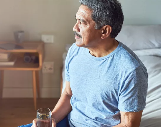 A Hispanic man sitting on the edge of his bed with a glass of water