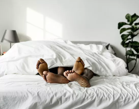 The feet of two Black people sticking out from under the covers of a large bed