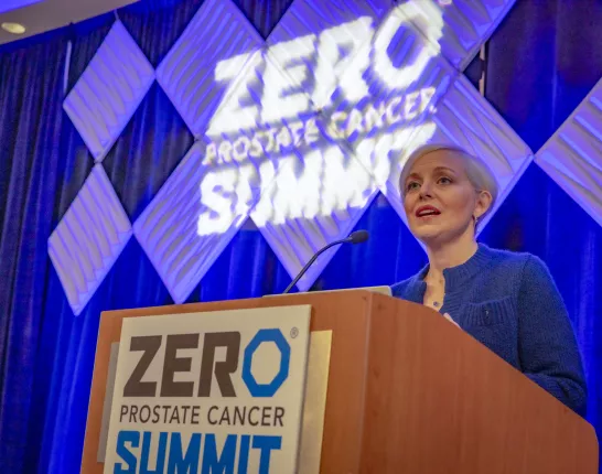 Blonde woman with very short hair standing behind a podium that has a sign saying "ZERO Prostate Cancer Summit"