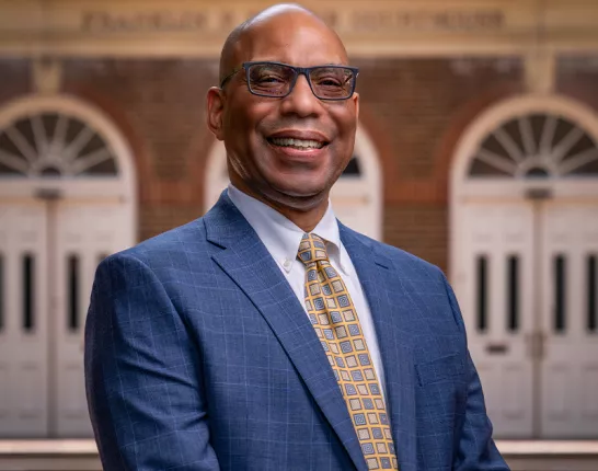 Brian Bragg, Chief Mission Officer, a Black man in a blue suit and yellow checkered tie standing in front of a brick building