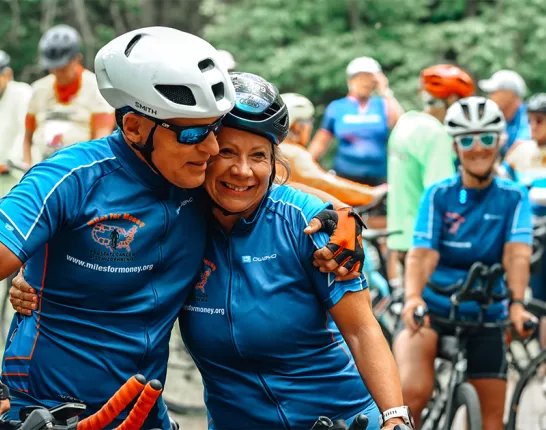 An older couple in cycling jerseys and helmets embracing while surround by many onlooking cyclists