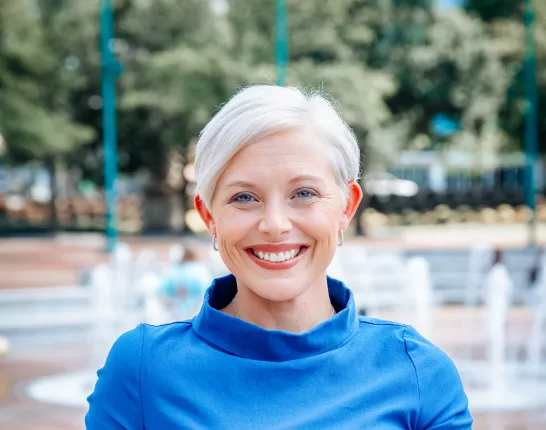 Blonde woman with short hair wearing a blue dress standing in front of a fountain