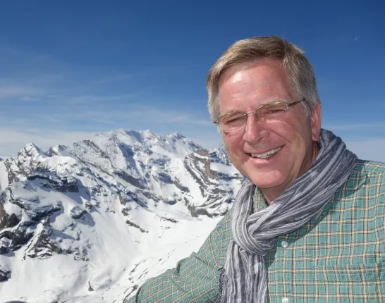 A blonde man with glasses standing in front of the Swiss Alps