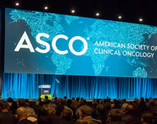 A large conference hall with a wide stage featuring a prominent display screen showing the ASCO (American Society of Clinical Oncology) logo and text in white against a turquoise world map background. The stage is framed by blue-lit curtains and lighting. The audience area is filled with attendees seated in rows, viewed from behind. The overall lighting creates a professional, dramatic atmosphere typical of major medical conferences.