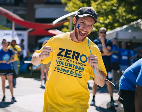 An energetic volunteer wearing a yellow ZERO Prostate Cancer Volunteer t-shirt with "I'm here to help" text smiles broadly at the camera on a sunny day. He has a blue ribbon painted on his face, with other volunteers visible in the background.