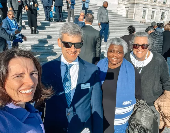 A group of people smiling on the steps of a large building, with some wearing blue scarves that say "ZERO Prostate Cancer." The setting appears to be a formal gathering or event.